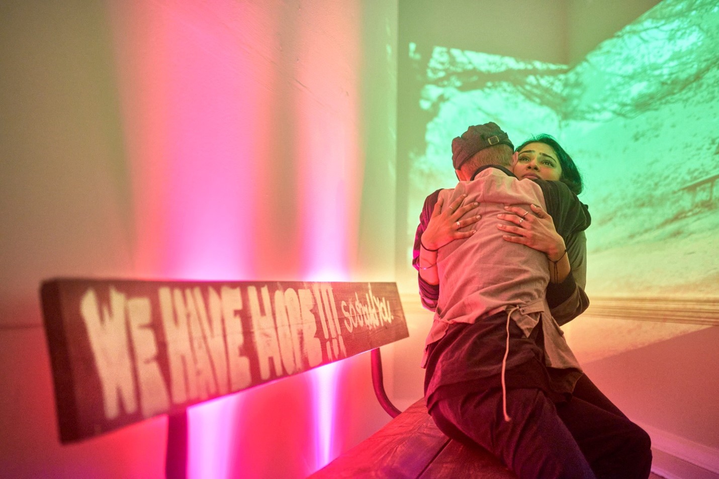 A photo of two people embracing on a bench. The bench and its surroundings are lit in bright pink and green.