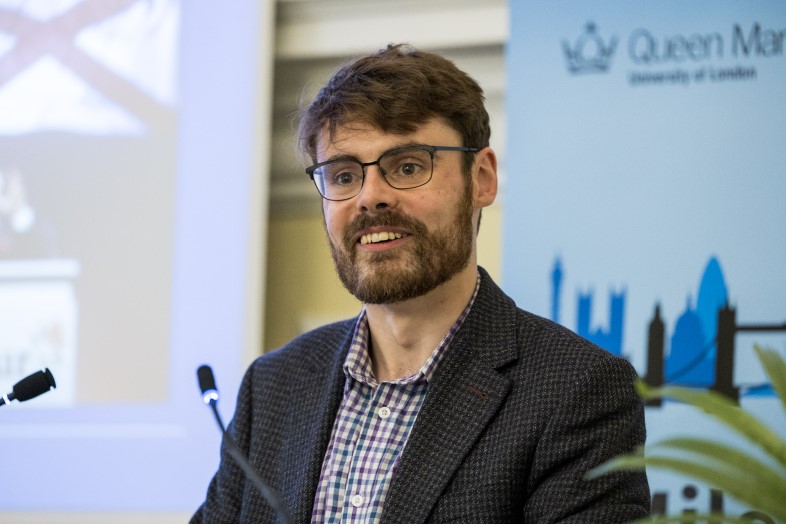 A photo of Colm Murphy speaking at a Mile End Institute event in front of a blue banner
