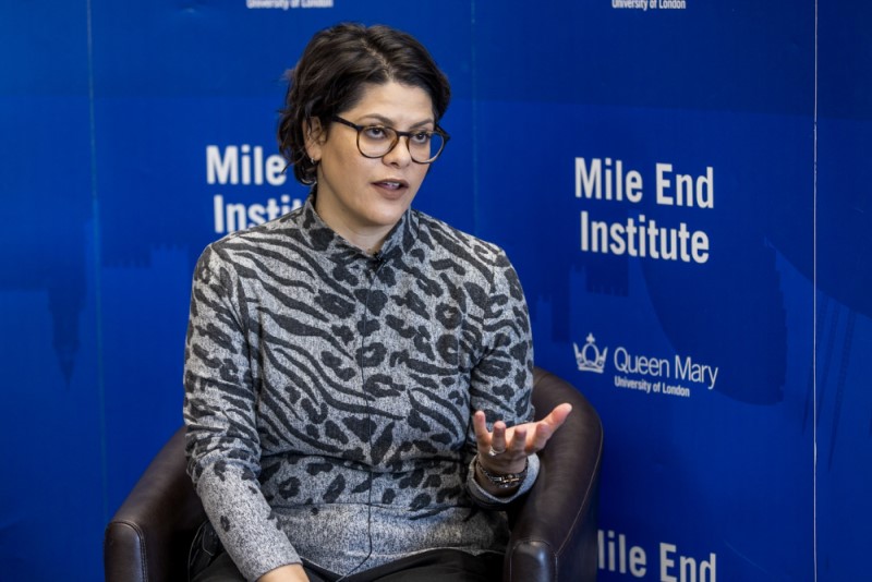 A photo of Sofia speaking at an event in front of a blue banner for the Mile End Institute