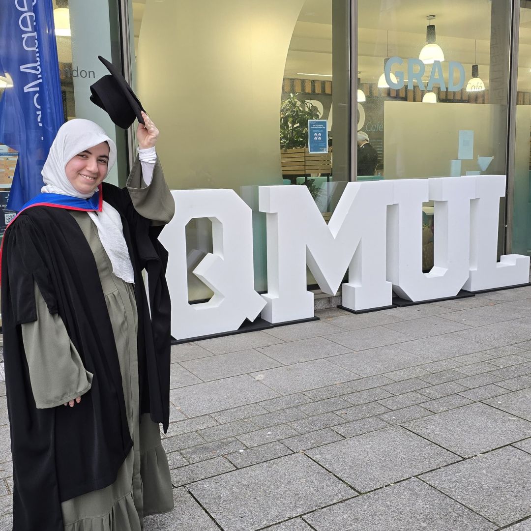 A student, dressed in graduation robes, stands in front of a large QMUL sign on graduation