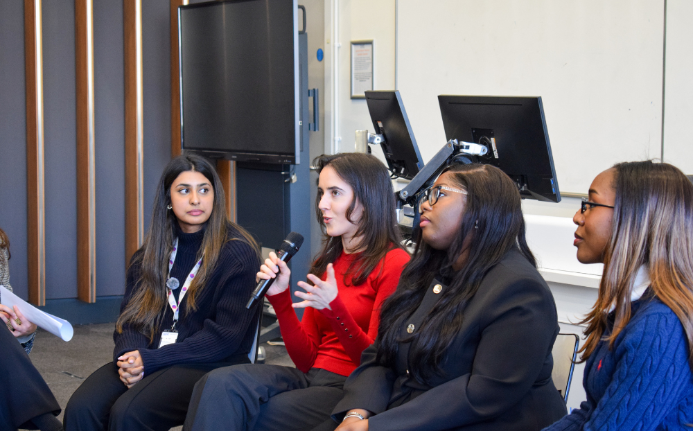 Image of 4 women sat on chairs in a lecture theatre in a panel and one holding a microphone