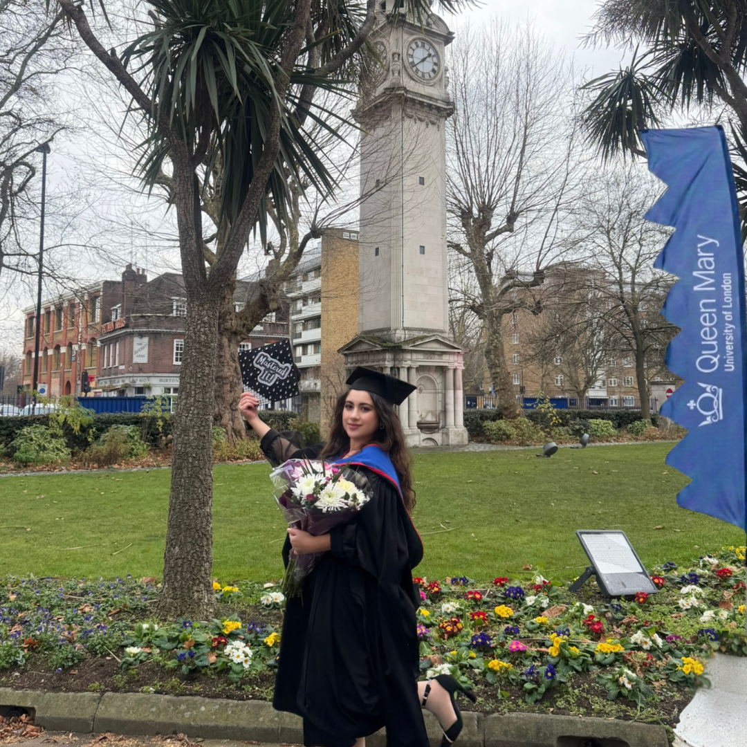 A student wearing a graduation gown carrying flowers and smiling infront of a clocktower at Queen Mary University of London