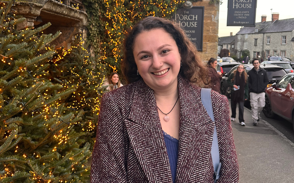 An image of a female student in smart casual clothes smiling in front of an inn and a christmas tree
