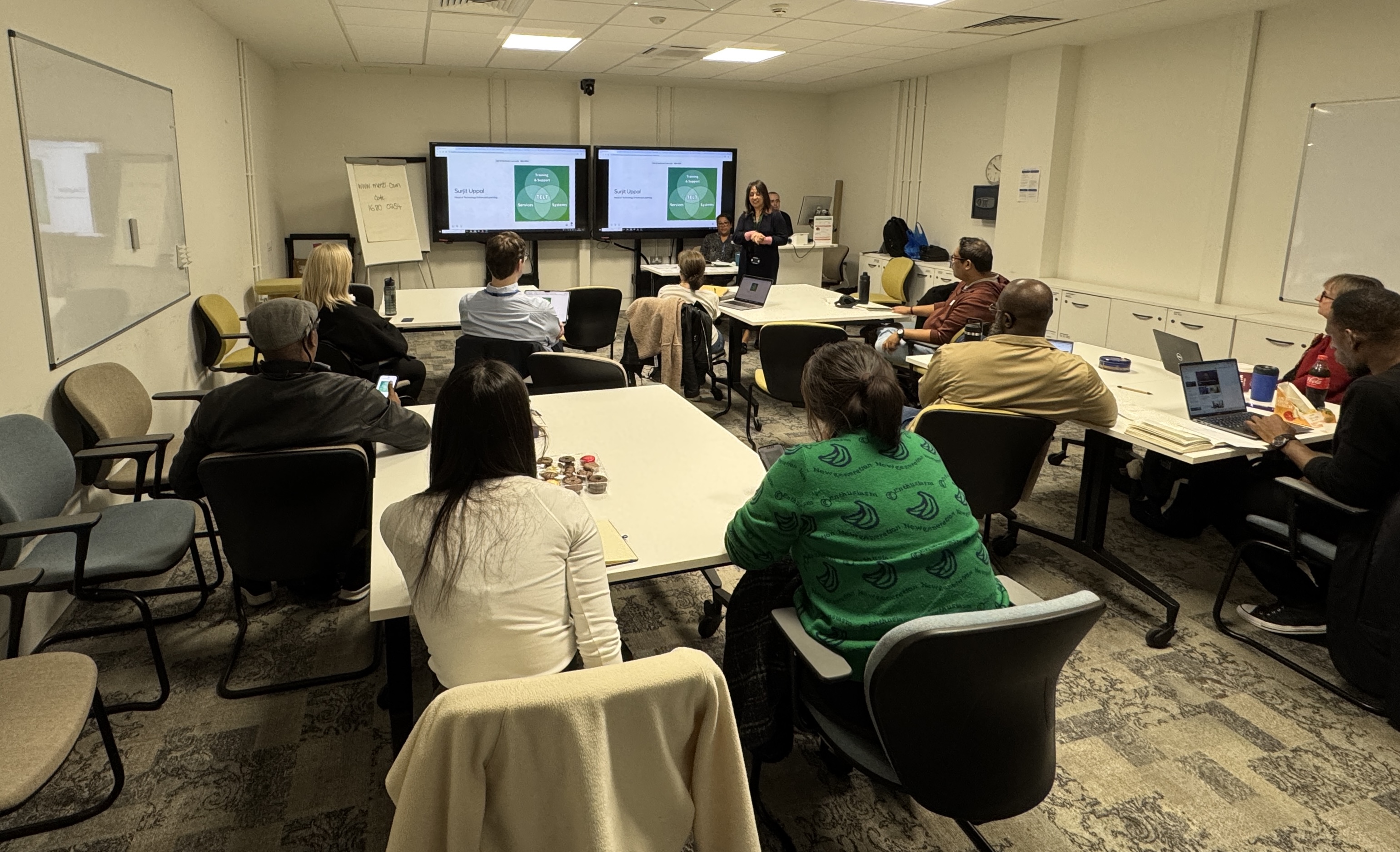 A training room with participants looking towards a presenter standing in front of multiple screens