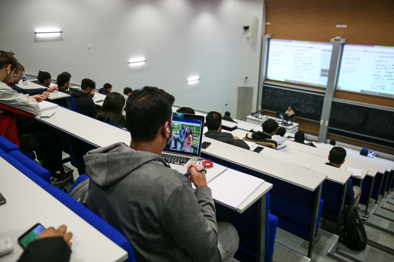 A student sitting at the back of a large tiered lecture theatre with a laptop on the table.  The lecturer is very far away.