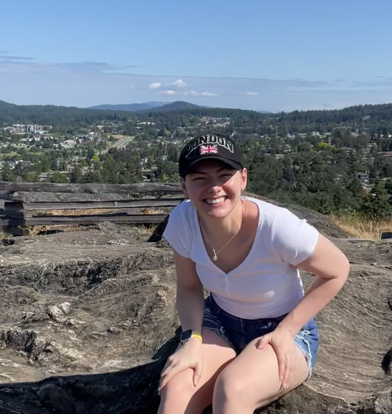Kailin Gillis sitting on a wall, looking at the camera smiling. There is a panoramic view in background with hills in the distance.