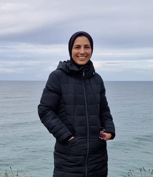 Meher Khan standing smiling at the camera. She is wearing all black and the sea is behind her.