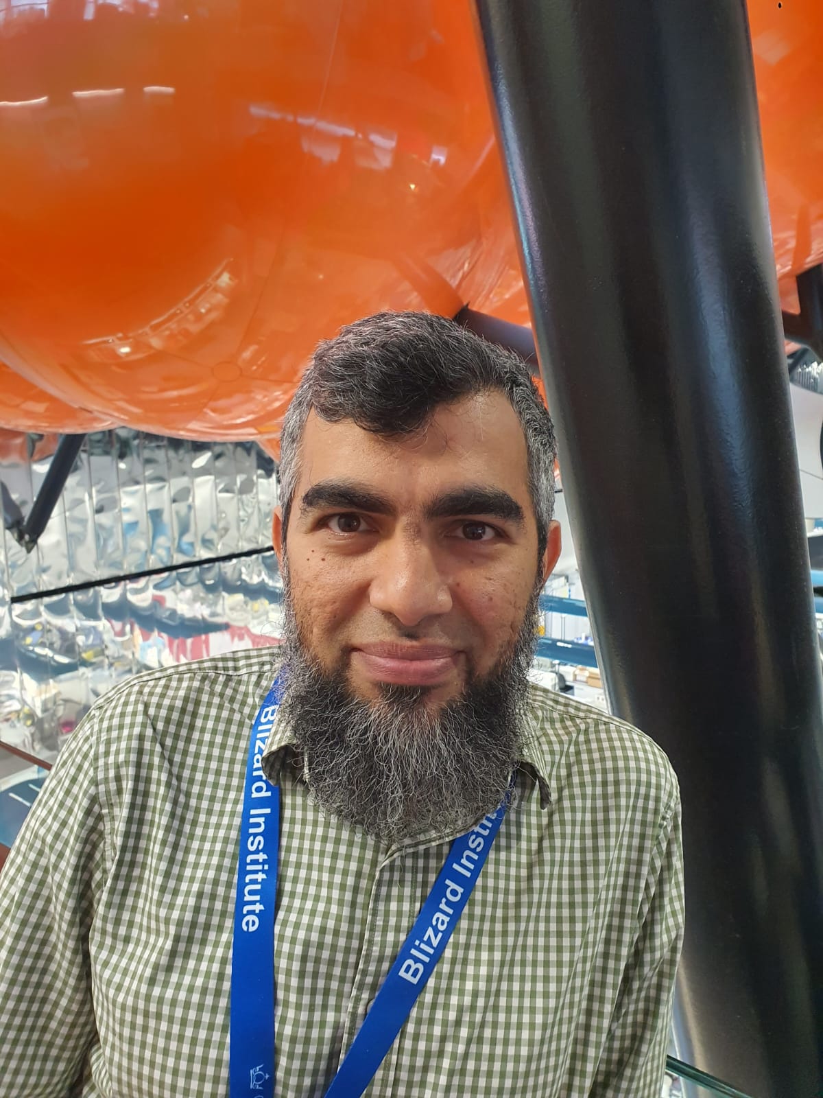 Atif Matin looking at camera wearing a shirt. Standing next to a black column with an orange background.