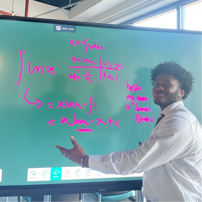 Samuel Ojeiwa TEL Student Ambassador. In a white shirt standing in front of a whiteboard with writing on it. Looking at camera while gesturing towards board.