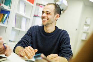 A man in a black top sitting at a table. Smiling with their head turned looking to the right. There is a shelving unit behind them.