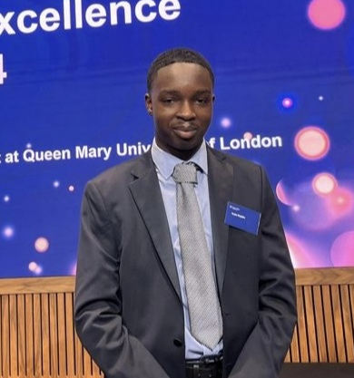Tosin looking at the camera, wearing a grey suit, shirt and tie. With a blue pocket square. Standing in front of a blue background.