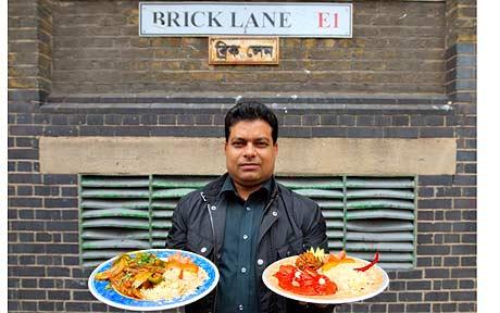 A man in a dark shirt holding two plates of food, looking at the camera, standing in front of a brick wall and a street sign.