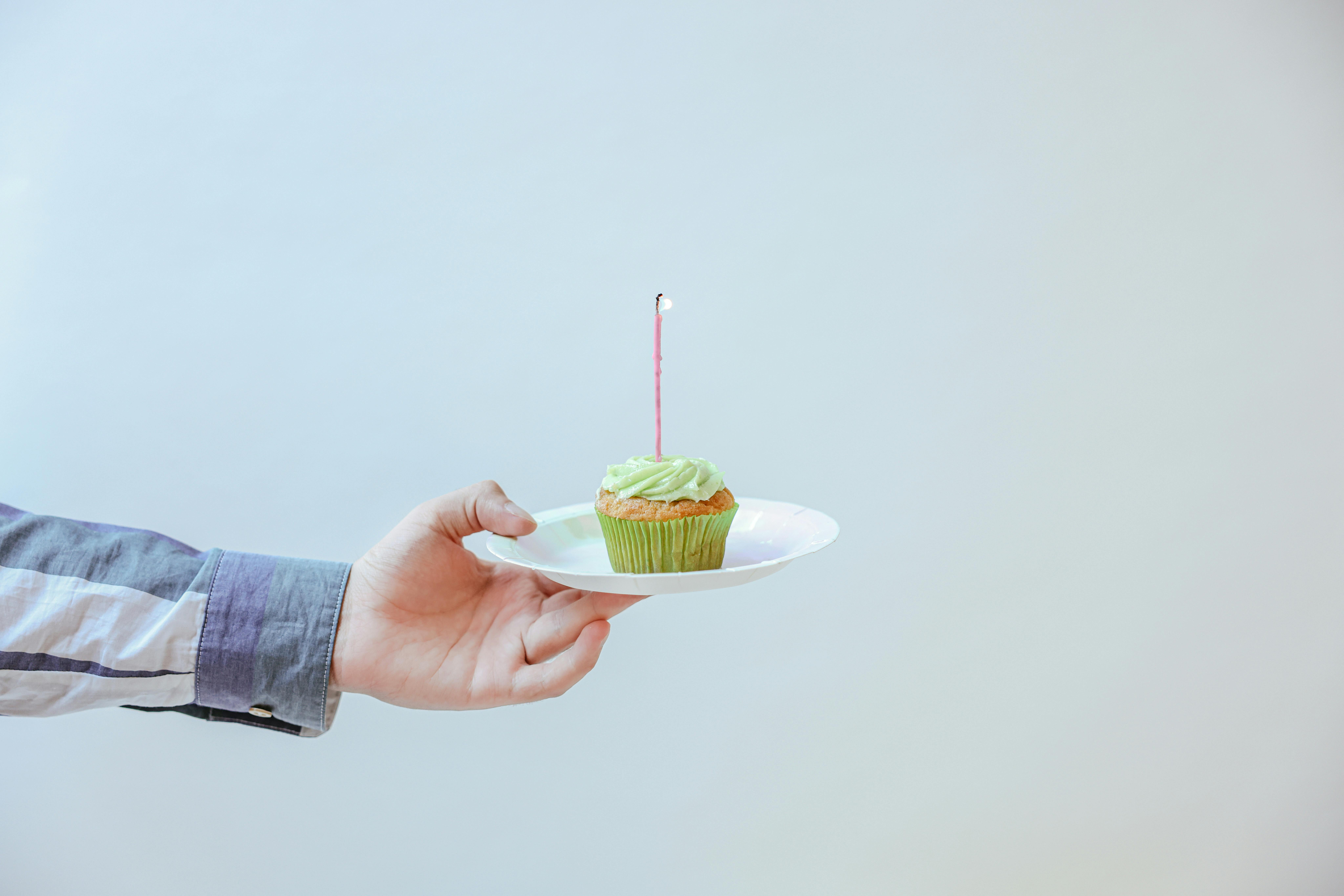 An extended arm in a shirt holding a cupcake with a candle on a small plate.