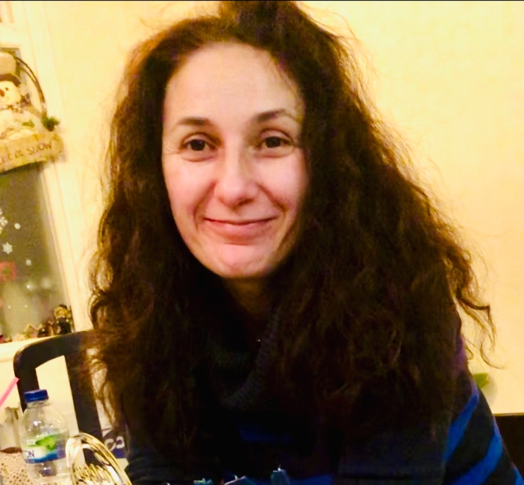 A photo of a lady smiling and looking towards the camera. She is wearing a dark top and has a yellow background behind.