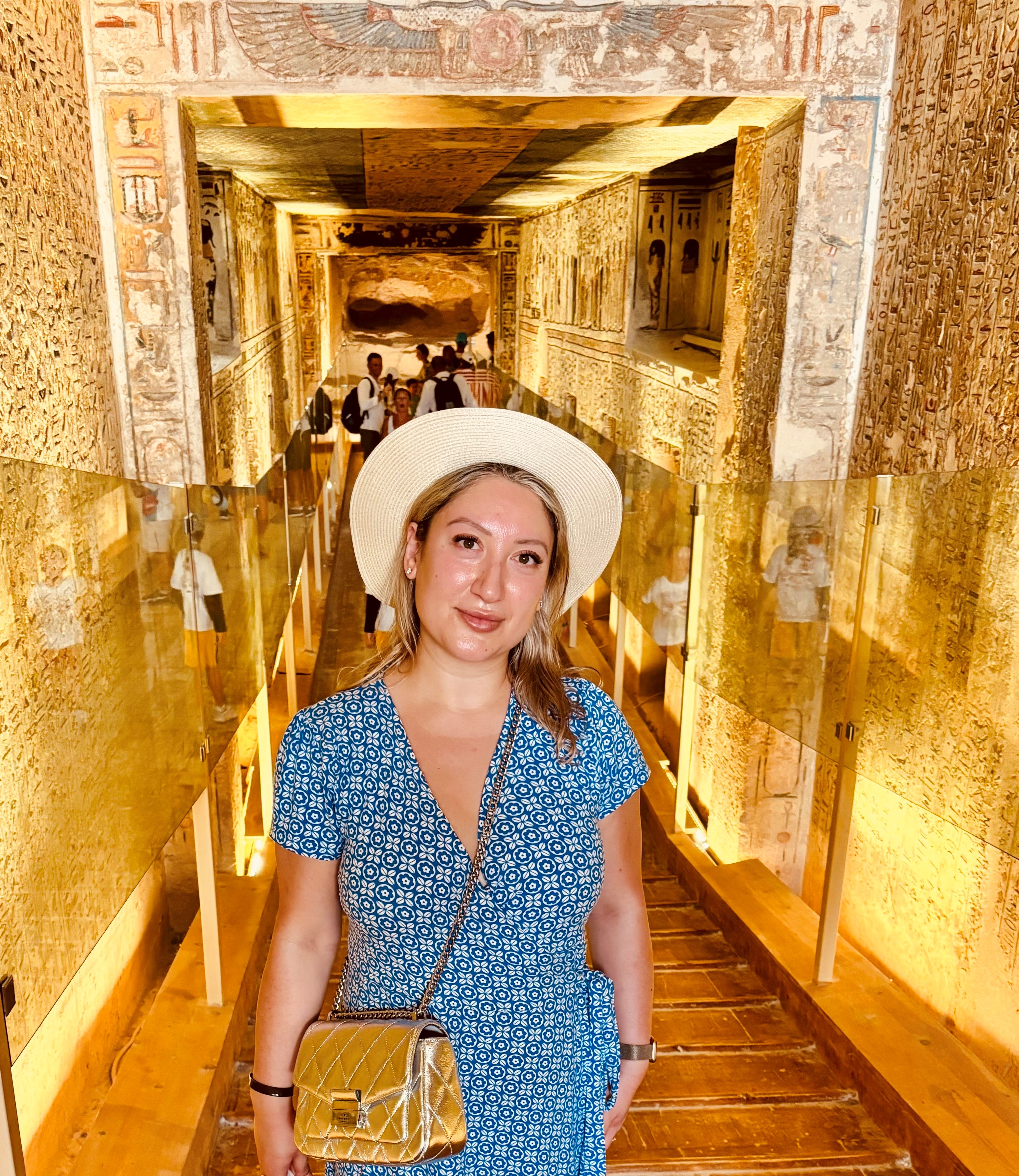 Yelena is standing in front of a wall covered in Egyptian hieroglyphs. She is wearing a blue dress and a white hat.